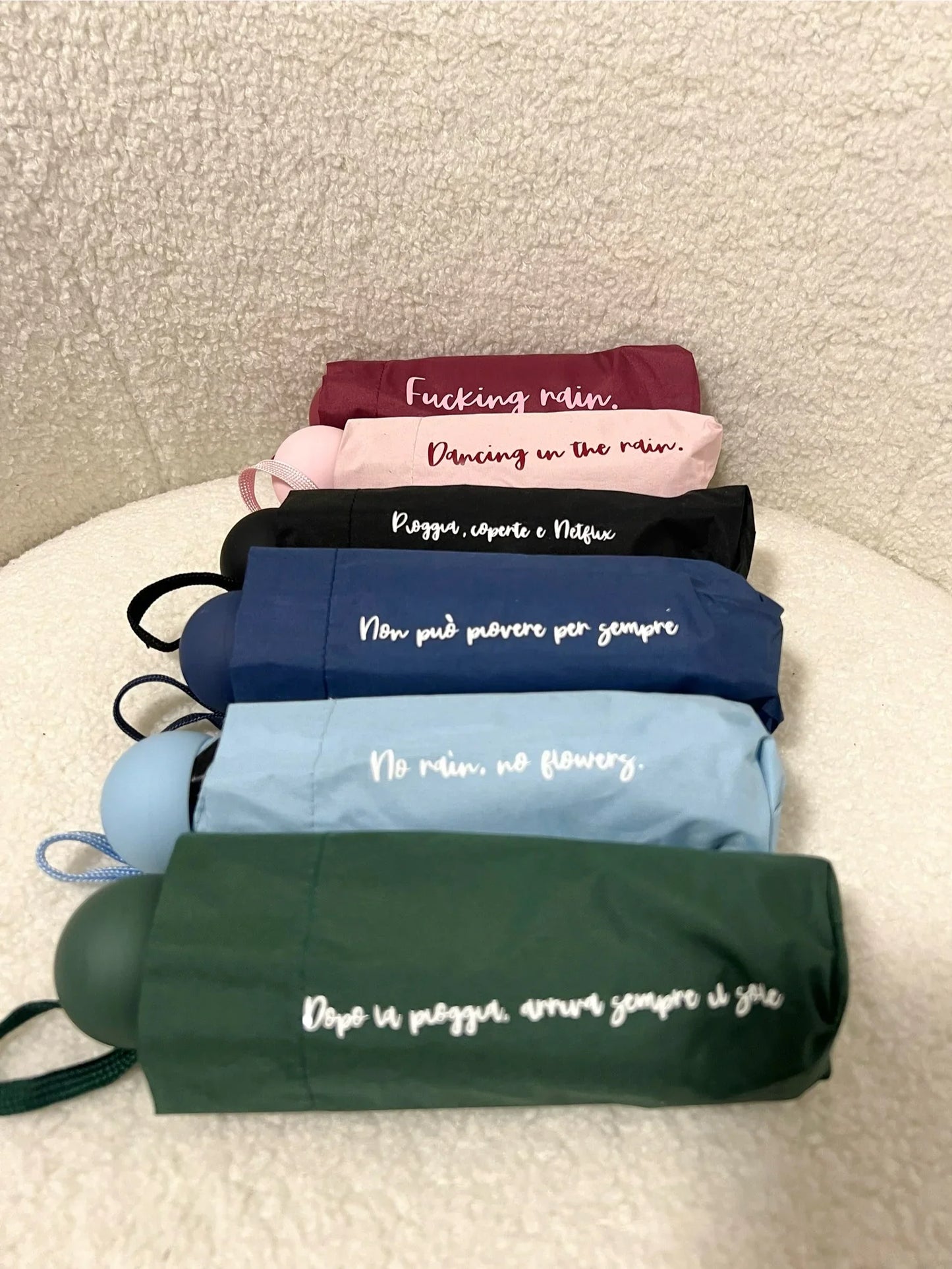 Sei ombrelli pieghevoli colorati con scritte ispirate alla pioggia, disposti su uno sfondo morbido