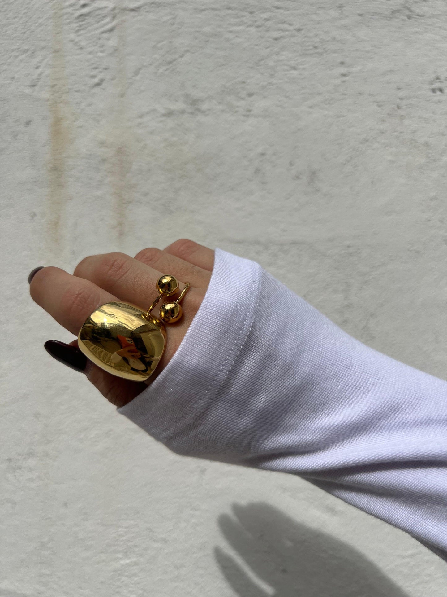 Mano con un anello dorato a forma di cuore e altro anello dorato con due sfere indossati sopra una maglia bianca a manica lunga