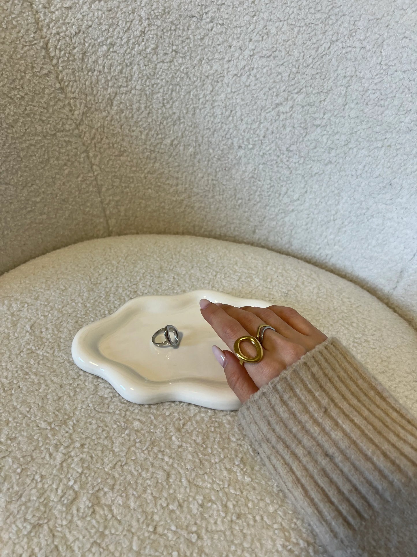 Mano con anelli dorati e argento posata su vassoio bianco ondulato su sedia beige in tessuto bouclé