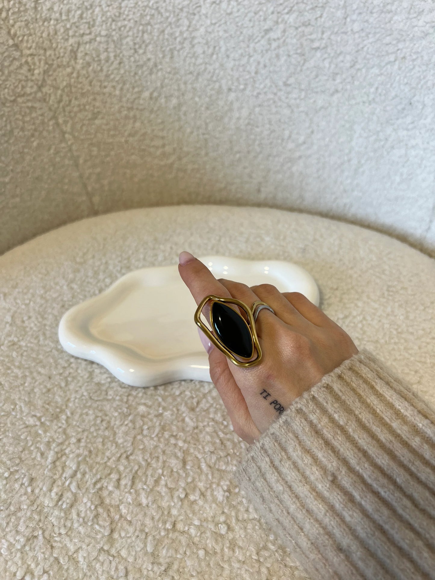 Mano con anello grande dorato con pietra nera ovale su tessuto beige e piattino bianco decorativo