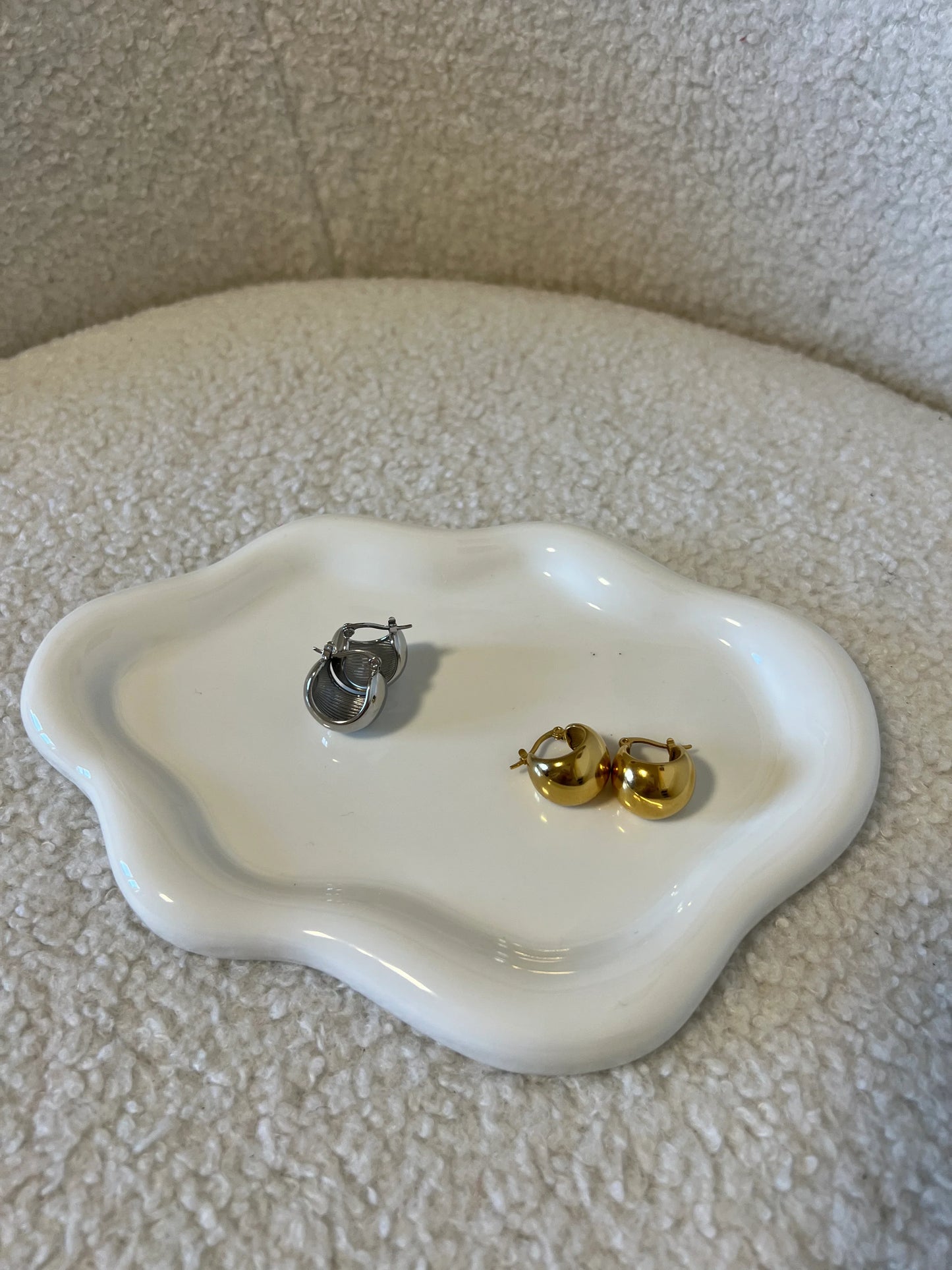 Coppia di orecchini a cerchio in oro e argento su piattino bianco ondulato, sfondo tessuto chiaro