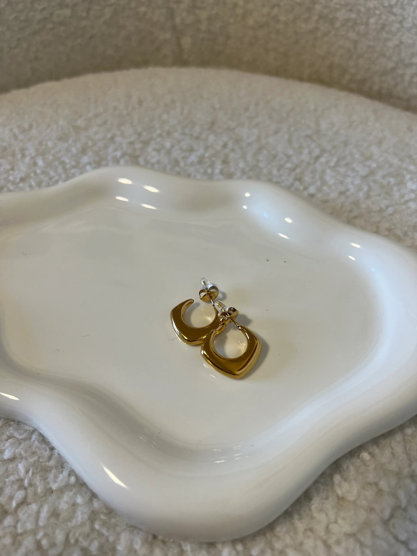 Orecchini a cerchio quadrato dorati su piatto bianco ondulato tessuto chiaro sullo sfondo
