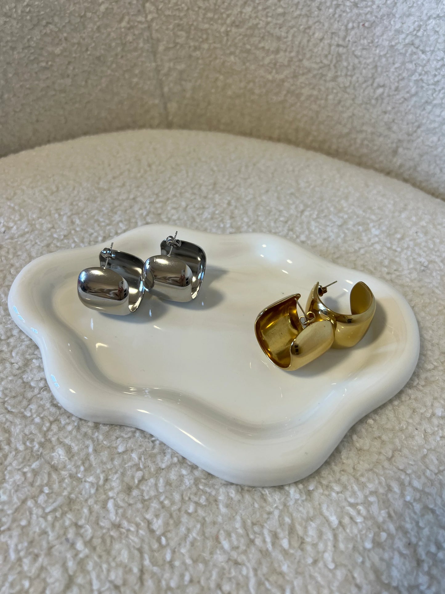 Orecchini a cerchio in metallo lucido argento e oro su piatto bianco ondulato su sfondo tessuto beige