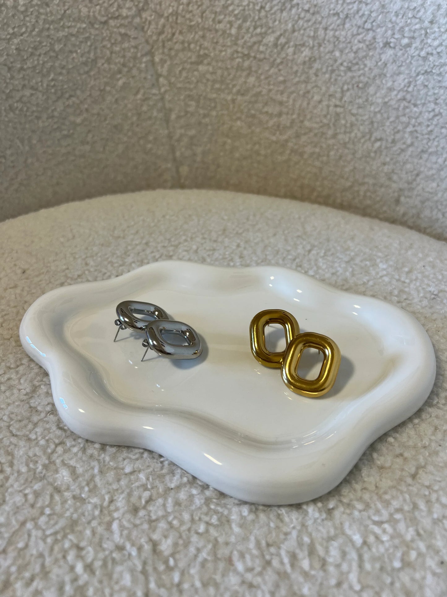 Orecchini Les Soeurs 33 in metallo oro e argento su piatto bianco con bordi ondulati, sfondo tessuto beige