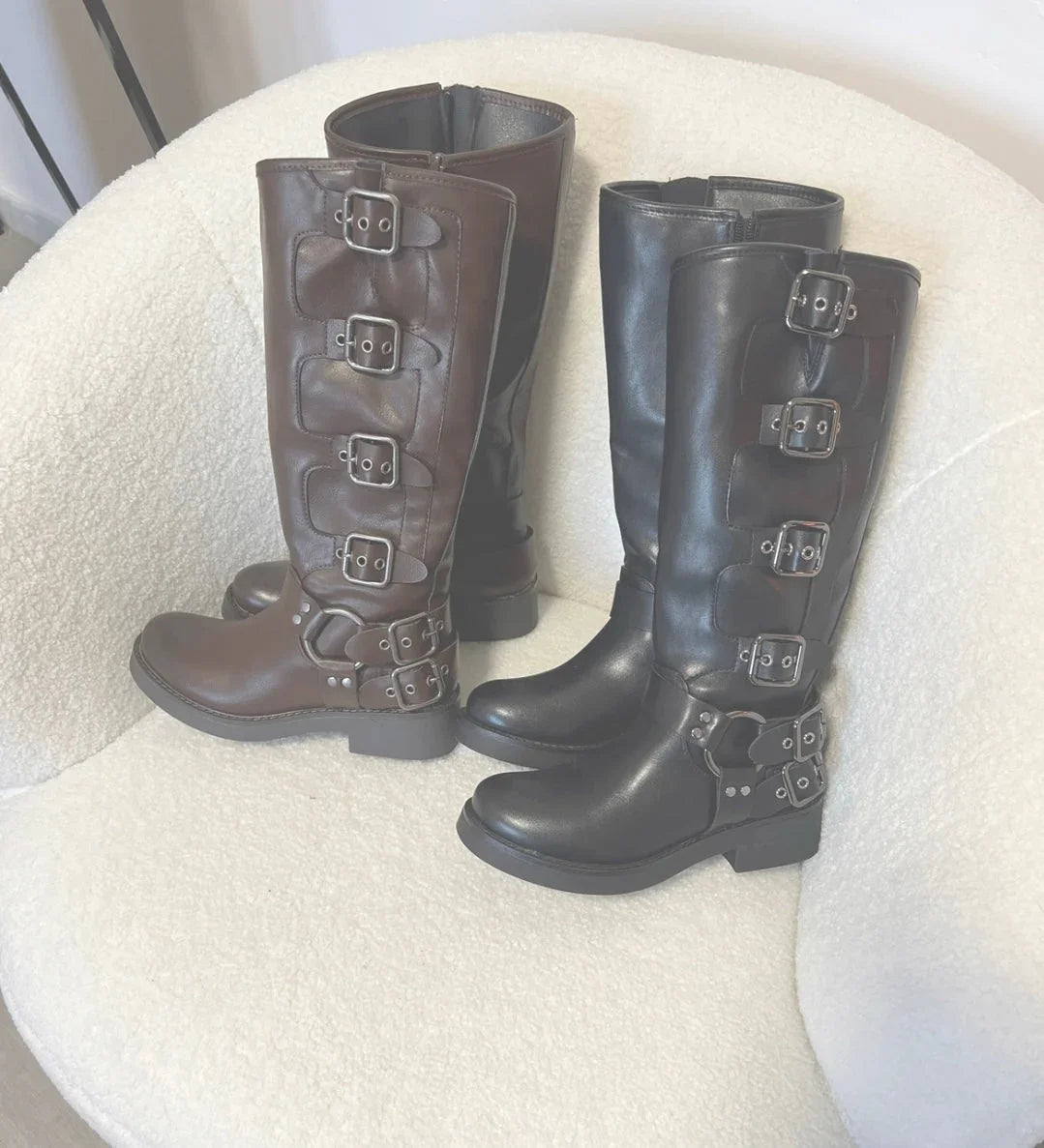 Stivali alti in pelle marrone e nera con fibbie metalliche, su poltrona bianca soffice